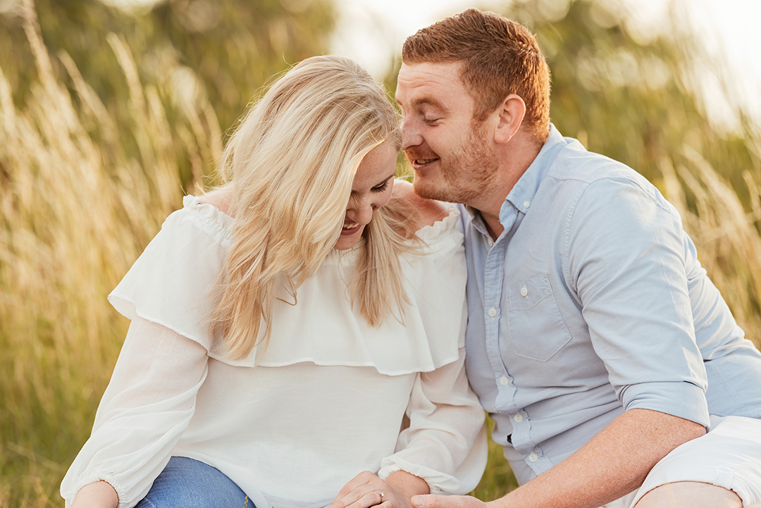 Couple sit in tall grass laughing Hylands Park engagement session