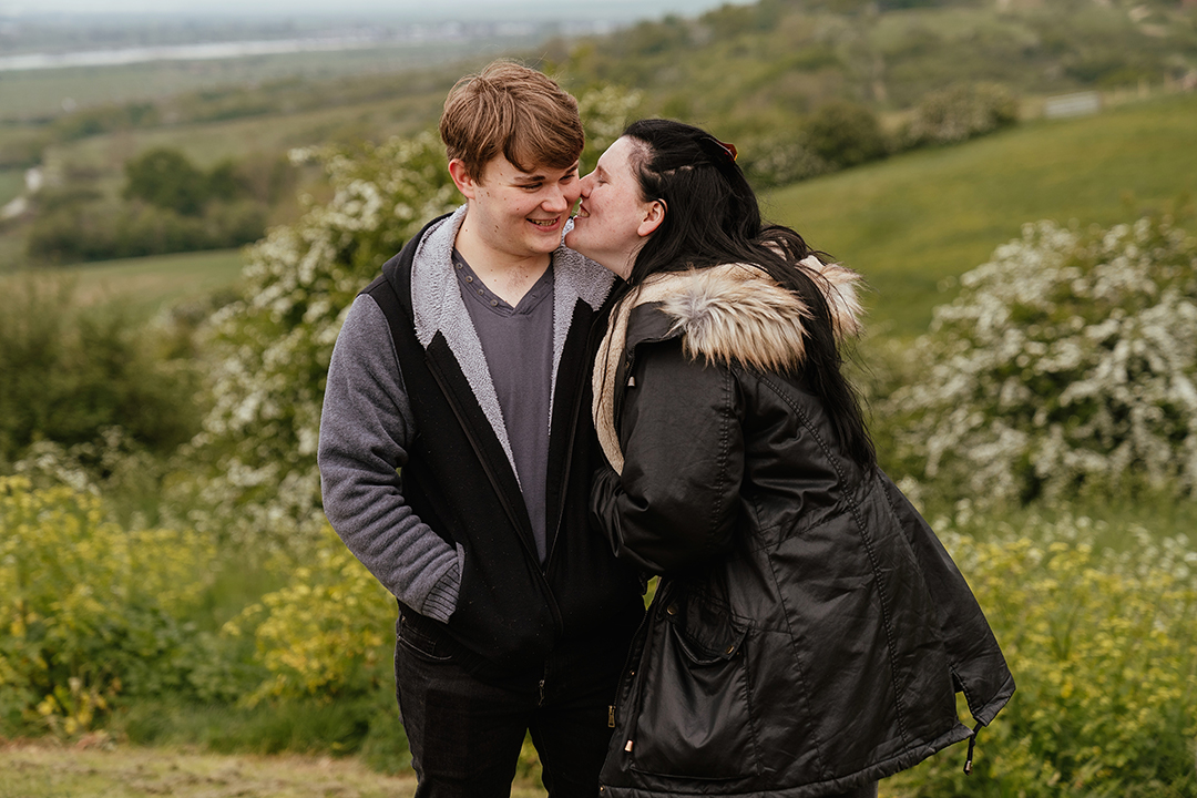 Couple stand with greenery in the background while she kisses his cheek Hadleigh Castle