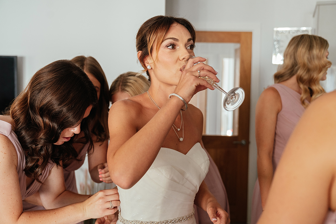Bride Drinks Prosecco While Bridesmaids Do Up Dress