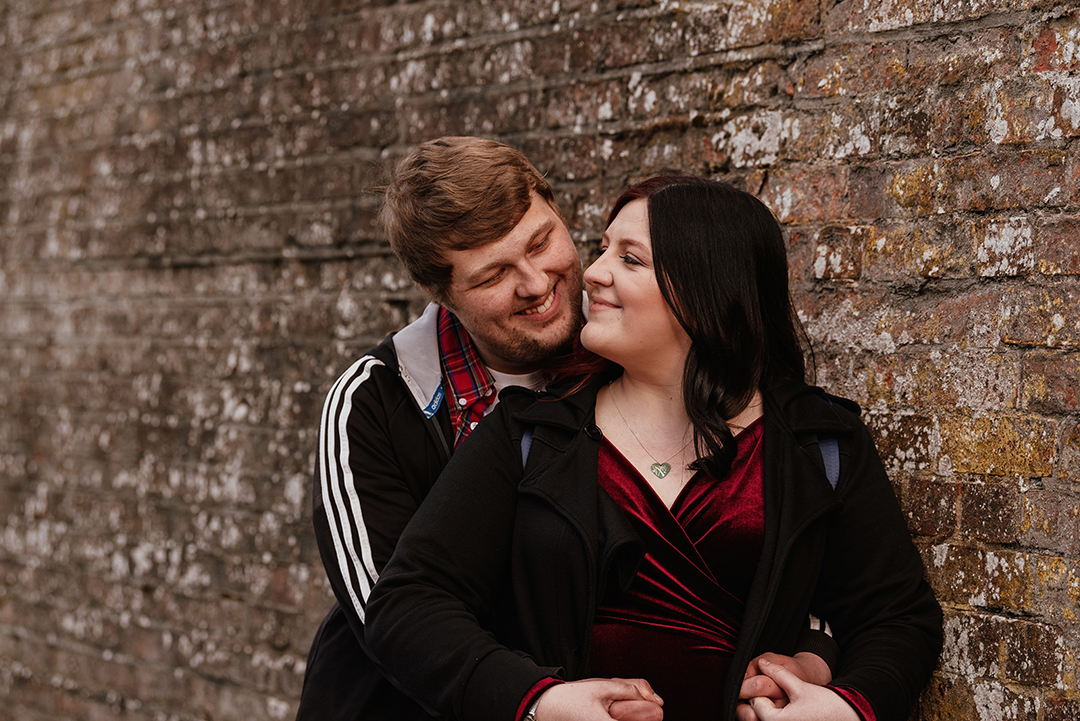 Couple lean again wall as he hugs her from behind looking to each other Castle Park Couples Shoot