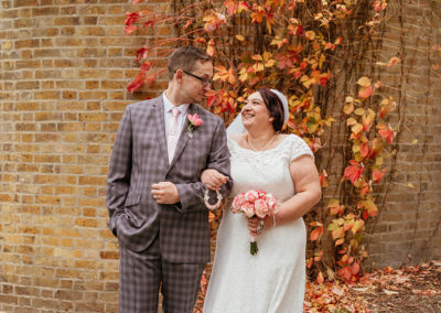 Couple look at each other smiling with autumn leaves on brick wall in background Moot House Wedding Photography