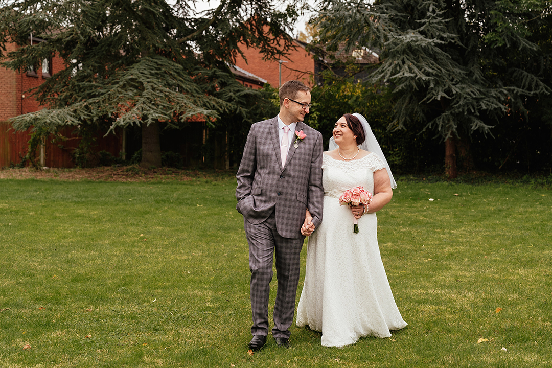 Couple walk towards camera with evergreen trees in background at Moot House Wedding Harlow