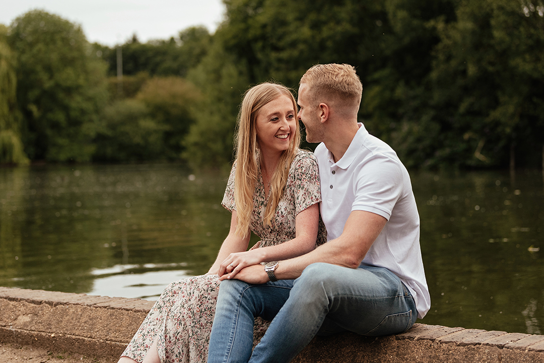 Couple sit looking at each other, on wall ledge by Lake Natural Pre Wedding Photoshoot at Stanborough Lake