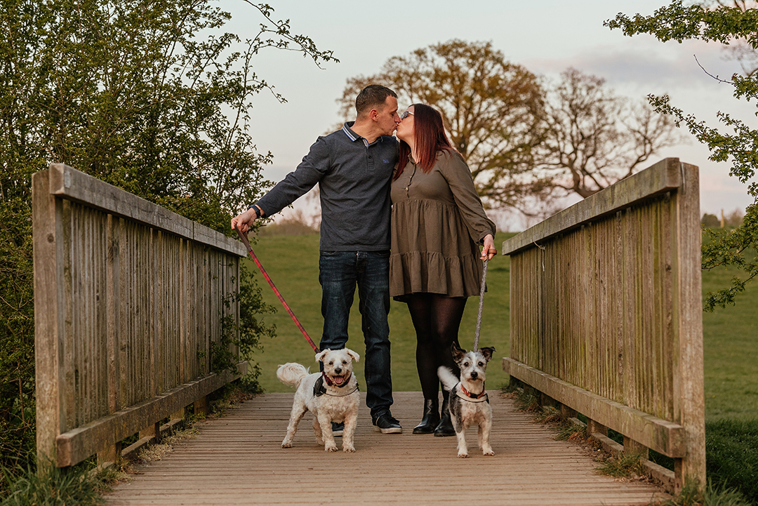 Couple stand kissing holding their dogs leads on bridge Pishiobury Park Couples Shoot with Dogs