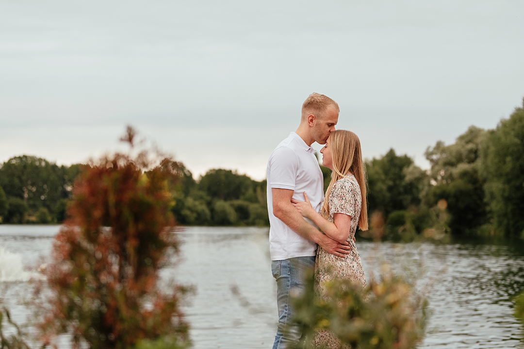 Couple behind the plants in front of the lake enjoying a private moment where he kisses her on the head at Stanborough Lake Couples Pre Wedding Photos