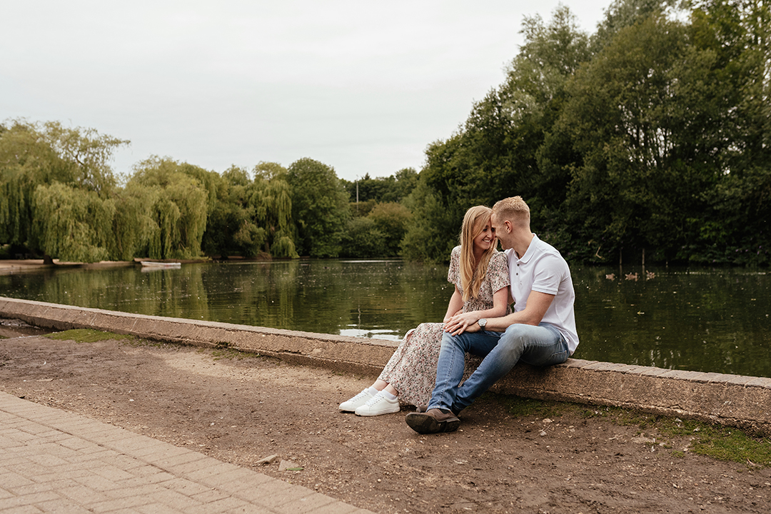 Full length image of couple sit looking at each other, on wall ledge by Lake Natural Pre Wedding Photoshoot at Stanborough Lake